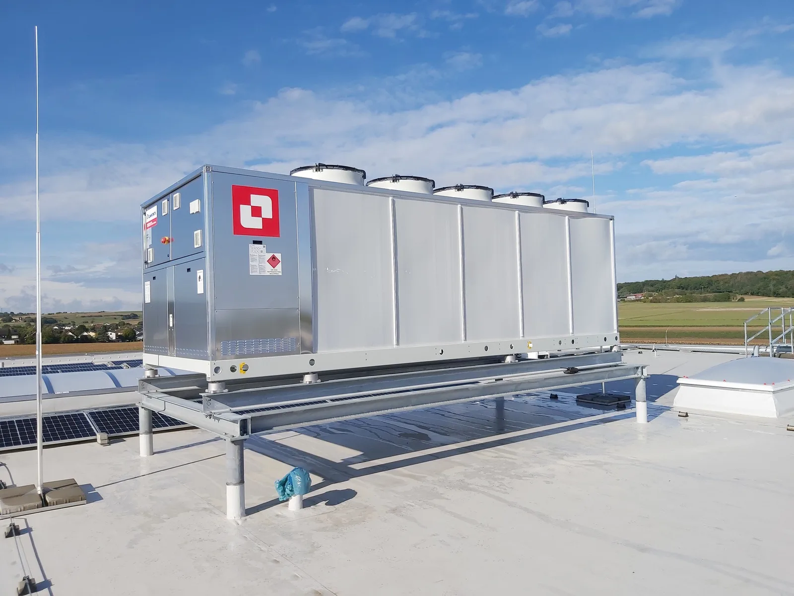 Rückkühlgerät auf einem Hallendach neben Solarmodulen, Blick über das Umland
