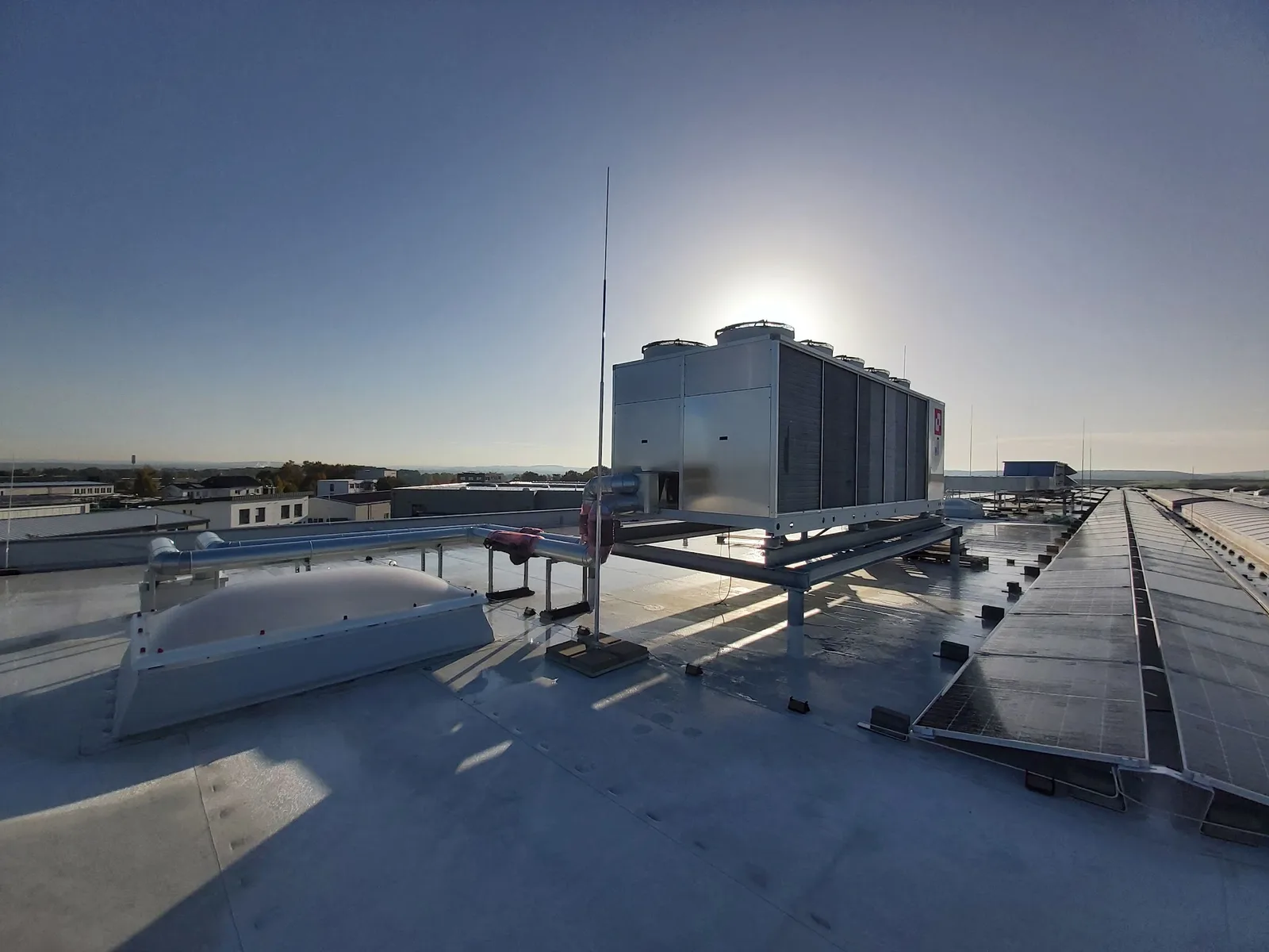 Großes Rückkühlgerät auf einem Flachdach mit Lüftungsrohren und Solarmodulen im Gegenlicht