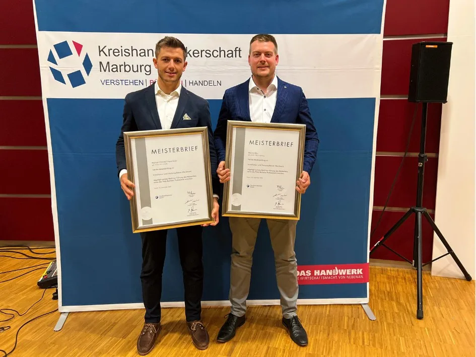 Henrik Dörr und Manuel Hasenauer mit ihren Meisterbriefen vor einer Wand der Kreishandwerkerschaft Marburg